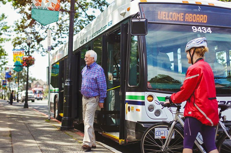 BC Transit - Senior man getting off a bus, woman on bike in foreground.